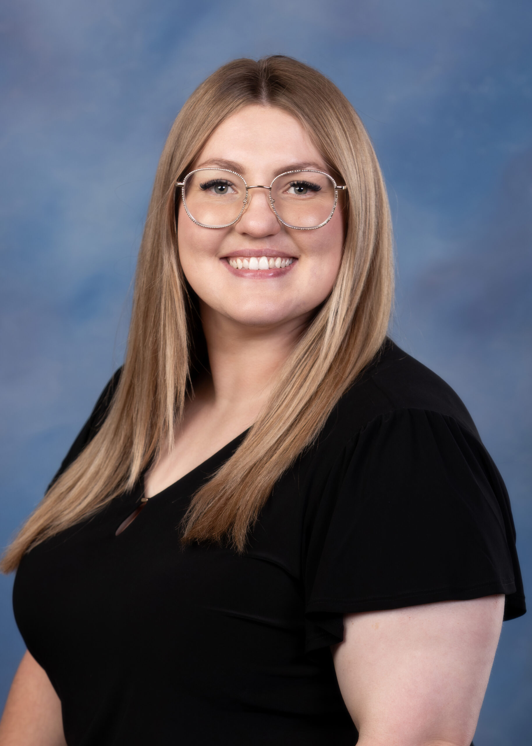 A white women with long blonde hair, she is smiling and is wearing a short sleeve black shirt