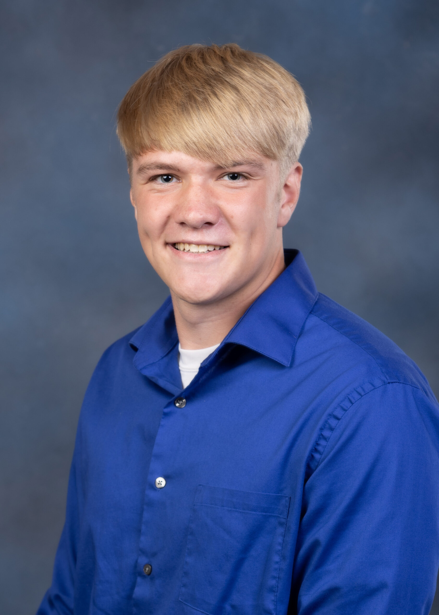 A white male with blonde hair, he is wearing a long sleeve blue shirt and smiling.