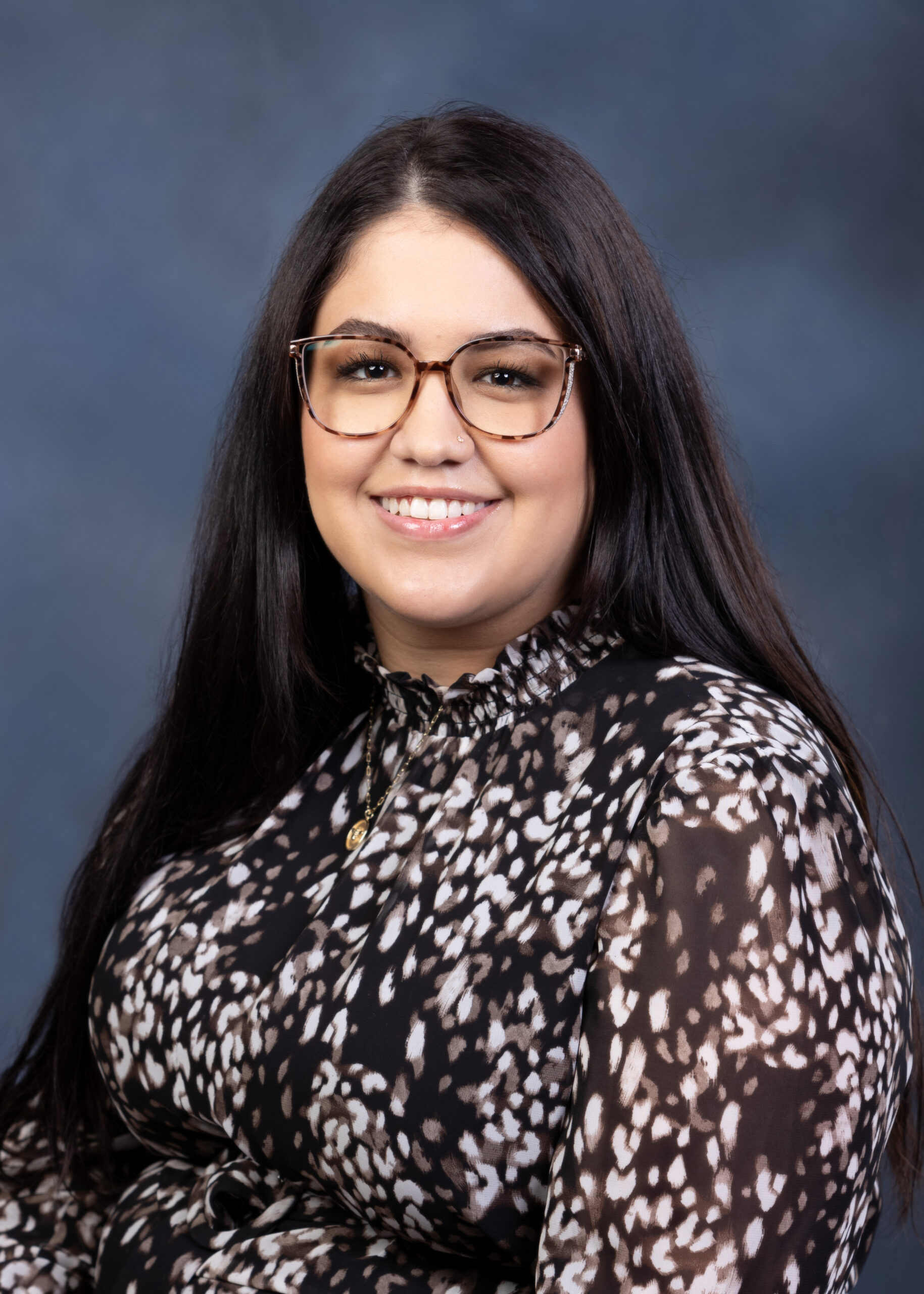 A white women with long brown hair, she has glasses on and is wearing a black shirt with white spots